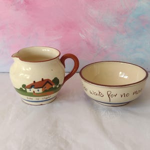 May include: A cream-coloured ceramic creamer and bowl set with brown rims. The creamer features a painted house and the text Isle of Wight. The bowl features the text Tide waits for no man written in black script along the outer rim.