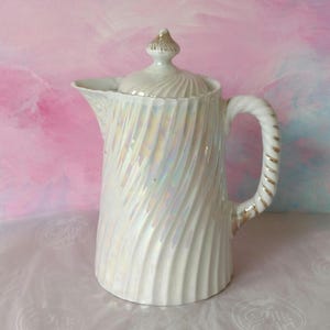 May include: A white ceramic jug with a lid, featuring a pearlescent finish and a spiral design. The handle has gold accents. The jug is designed for serving beverages.