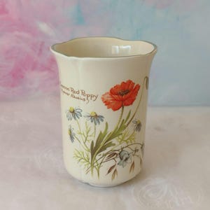 May include: Cream ceramic vase with a wavy rim and a floral design. The design features a red poppy, blue daisies, and green leaves. Text on the vase reads "Common Red Poppy, Papaver Rhoeas."