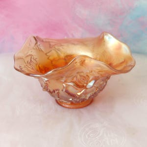 May include: An orange iridescent glass bowl with a ruffled edge. The bowl features a textured surface with floral designs and a circular base. The glass reflects light, producing a shimmering effect. Likely a decorative item.