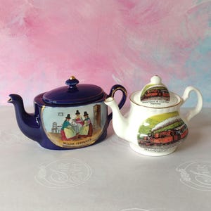 May include: A dark blue teapot featuring a central illustration of three figures in traditional Welsh costumes and a white teapot decorated with a steam train graphic labelled Welshpool & Llanfair Railway with gold trim on both lids and handles.