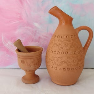 May include: A terracotta pitcher and mortar set. The pitcher has a handle and a spout, with floral designs and a series of small holes. The mortar has a pedestal base and a pestle. The set is a warm, earthy tone.
