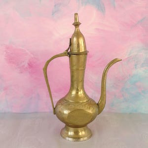 May include: A vintage brass jug with a long spout, curved handle, and decorative patterns. The jug has a rounded body and a tall, conical lid. The background is a blend of pink and blue hues.