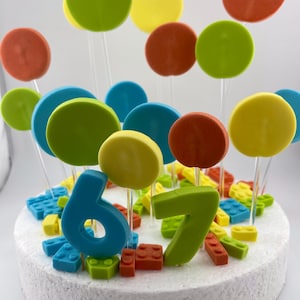 Könnte beinhalten: Ein bunter Geburtstagskuchen mit weißer Basis, verziert mit blauen und grünen Zahlen 6 und 7, bunten runden Toppern und Spielzeugstein-Akzenten. Der Kuchen hat orange, gelbe, grüne und blaue Elemente und wirkt festlich.