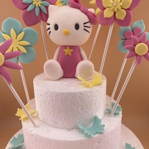 Puede incluir: Un pastel blanco de dos niveles decorado con una figura de Hello Kitty y flores coloridas. El pastel está adornado con flores rosas, amarillas y azules sobre palitos y detalles de mariposas. La figura de Hello Kitty lleva un vestido y un lazo rosas.