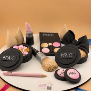May include: A collection of edible cake toppers shaped like makeup items, including a black MAC eyeshadow palette, a makeup brush, a lipstick, two beige sponges, a black bow, and several round compacts with the MAC logo in pink and black colors.