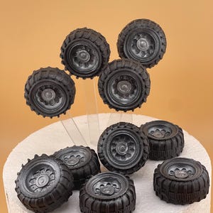 May include: A selection of small, black tyre decorations, possibly for a cake or craft. The tyres feature detailed tread patterns and silver-coloured wheel centres. Some are on clear supports, others rest on a white, textured surface.