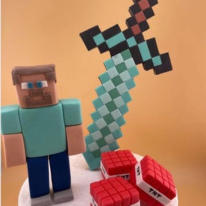 May include: A Minecraft-themed cake topper set featuring a pixelated character, a sword, and TNT blocks. The character has a teal shirt and blue pants. The sword is light blue, black, and red. The TNT blocks are red with white bands and the text "TNT".