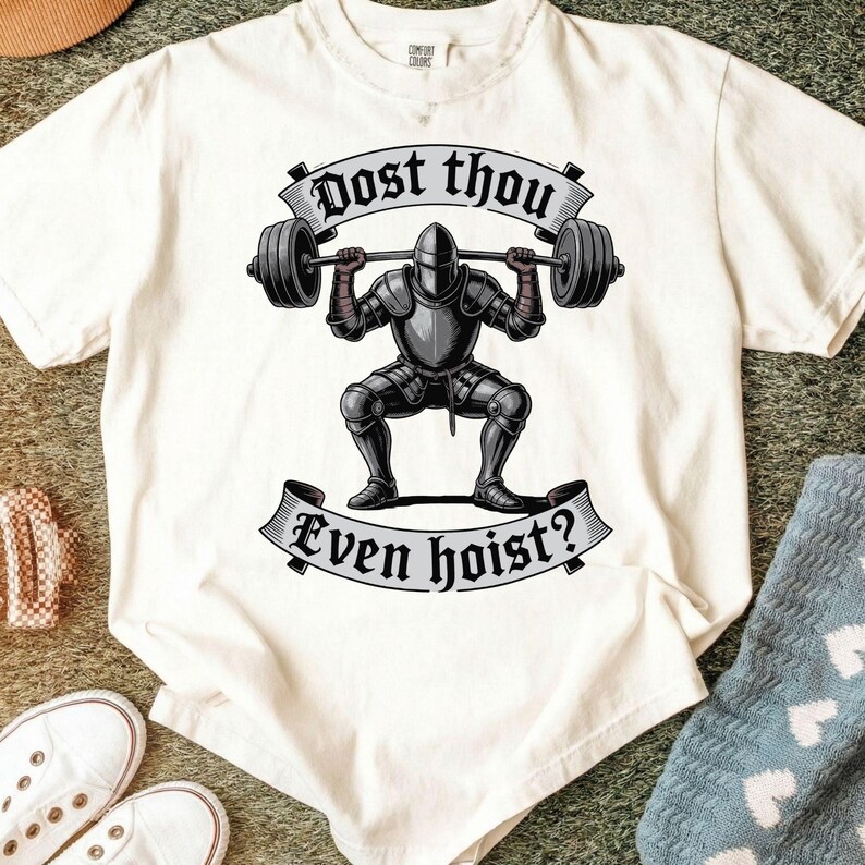 May include: Off-white t-shirt featuring a weightlifting knight graphic. The knight, clad in armor, is squatting while lifting a barbell. The text "Dost thou Even hoist?" is displayed above and below the knight in a banner.