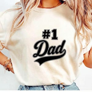 May include: A cream-colored short-sleeve t-shirt featuring the black text #1 Dad printed on the front. The design includes the number 1 in a block font above the word Dad written in a bold, cursive script with a decorative underline flourish.
