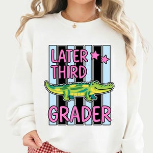 Könnte beinhalten: Weißes Sweatshirt mit dem Text "Later Third Grader" in Rosa, mit einer Illustration eines grünen und gelben Alligators. Das Design umfasst rosa Sterne und einen Hintergrund aus schwarzen und hellblauen Streifen.