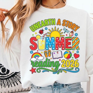 Könnte beinhalten: Weißes Sweatshirt mit farbenfrohem Grafikdesign. Das Design enthält den Text "UNEARTH A STORY SUMMER READING 2026" mit Illustrationen von Sonne, Regenbogen und sommerlichen Symbolen. Das Sweatshirt ist ein lässiger Stil.