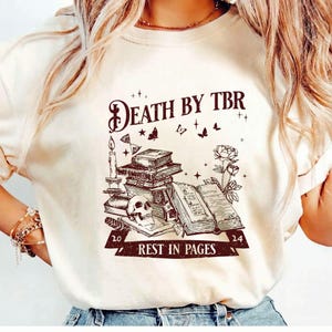 Może przedstawiać: Kremowa koszulka z nadrukiem graficznym. Projekt zawiera napis "DEATH BY TBR" nad stos książek, czaszką i świecą. Poniżej znajduje się fraza "REST IN PAGES" z liczbami "20" i "24".