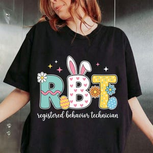 Könnte beinhalten: Schwarzes T-Shirt mit einem farbenfrohen Grafikdesign mit den Buchstaben "RBT", die mit Ostermotiven verziert sind. Das Design umfasst Hasenohren, Blumen und Ostereier. Darunter steht der Text "registered behavior technician".
