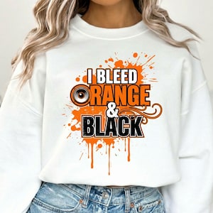 以下が含まれることがあります： オレンジと黒で「I Bleed Orange & Black」の文字が入った白いスウェットシャツ。デザインにはオレンジ色のスプラッシュ効果とスピーカーのグラフィックが含まれています。カジュアルで快適なトップス。