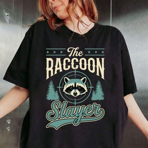 May include: Black t-shirt with a raccoon graphic inside a target reticle, flanked by pine trees. The text reads "The Raccoon Slayer" in a vintage style font. The shirt is a crew neck.