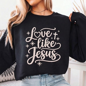 Peut inclure: Sweat-shirt noir à manches longues avec l'inscription blanche "Love like Jesus". Le design comprend un cœur, une croix et des étoiles. Le sweat-shirt est en matière douce.