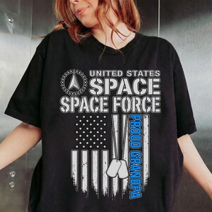 Op de afbeelding: Zwart T-shirt met de tekst "UNITED STATES SPACE FORCE" en een versleten Amerikaans vlagontwerp. Inclusief hondenpenningen en de woorden "PROUD GRANDPA" in blauw. Het shirt heeft een ronde hals.