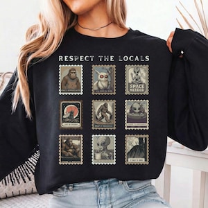 Pode incluir: Camisola preta de manga comprida com o texto "RESPECT THE LOCALS" acima de nove desenhos de selos postais. Os selos apresentam ilustrações de criptídeos e alienígenas com frases como "SPACE APPRECIATED" e "SPACE NEEDED".