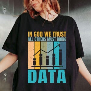 Può includere: T-shirt nera con la scritta "IN GOD WE TRUST ALL OTHERS MUST BRING DATA." Il design presenta un grafico con barre e linee ascendenti nelle tonalità dell'arancione, giallo, verde e blu. Il testo è in grassetto, senza grazie.