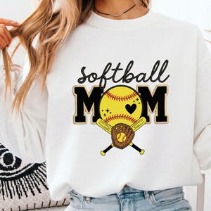 Peut inclure: Sweat-shirt blanc avec l'inscription "softball MOM" en noir et or. Le "O" de MOM est une balle de softball jaune avec un cœur noir, des battes croisées et un gant marron.