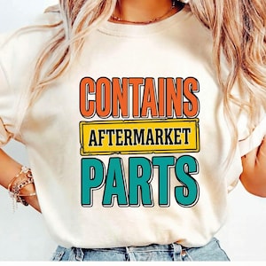 Puede incluir: Camiseta color crema con el texto "CONTAINS AFTERMARKET PARTS" en naranja, amarillo y turquesa. El texto está en negrita, con una fuente retro. La camiseta es de estilo casual.