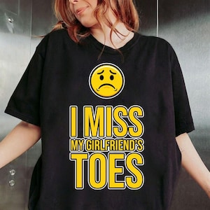 May include: Black t-shirt with the text "I MISS MY GIRLFRIEND'S TOES" in yellow, outlined in white. Above the text is a yellow sad face emoji. The shirt is a casual style.