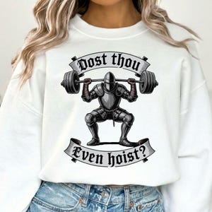 May include: White sweatshirt featuring a graphic of a knight in full armor lifting a barbell. The text "Dost thou" is above the knight, and "Even hoist?" is below. The knight is in a squat position, with the barbell overhead.