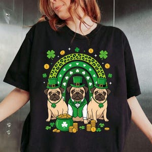 May include: Black t-shirt featuring three pugs dressed for St. Patrick's Day. The pugs wear green hats and bow ties, with a green rainbow and gold coins in the background. A pot of gold and shamrocks complete the festive design.