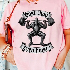 May include: Pink t-shirt featuring a graphic of a knight in armor lifting a barbell. The text on the shirt reads "Dost thou even hoist?" in a banner design. The knight is in a squat position.