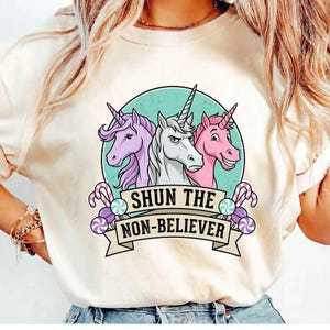 Op de afbeelding: Crèmekleurig T-shirt met een afbeelding van drie eenhoorns in een turquoise cirkel, met de tekst "SHUN THE NON-BELIEVER" op een banner. De eenhoorns zijn paars, wit en roze, met snoepaccenten.
