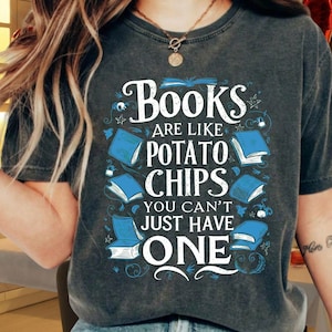 Puede incluir: Camiseta gris oscuro con la frase "Books are like potato chips, you can't just have one." El diseño incluye ilustraciones de libros azules. Una camiseta informal para amantes de los libros.