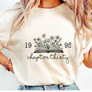 May include: Cream-colored t-shirt featuring a black graphic of an open book with flowers and the text "1995 chapter thirty". The shirt has short sleeves and a relaxed fit, suitable for casual wear.