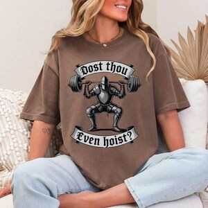 May include: Brown t-shirt featuring a graphic of a knight lifting a barbell. The text on the shirt reads "Dost thou even hoist?" in a banner design. The knight is in full armor.