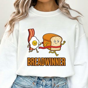 Op de afbeelding: Witte sweatshirt met een cartoonafbeelding van een brood dat naar een rode finishlijn rent, met een gebakken ei en spek in de achtervolging. Het woord "BREADWINNER" is in oranje blokletters gedrukt.