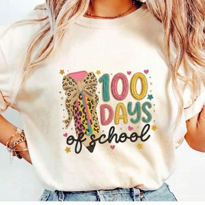 Può includere: T-shirt color crema con una grafica colorata che celebra i 100 giorni di scuola. Il design include una matita con un fiocco, la scritta "100 DAYS of school", cuori e stelle. La matita ha un motivo leopardato.