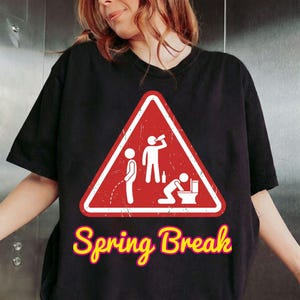 May include: Black t-shirt with a red warning sign graphic depicting stick figures drinking, urinating, and vomiting. The words "Spring Break" are written in yellow with a pink outline below the graphic.