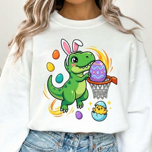 May include: White sweatshirt featuring a cartoon dinosaur wearing bunny ears, holding a purple Easter egg, and playing basketball. Colorful eggs and a hatching chick are also shown. The design has a playful, festive theme.