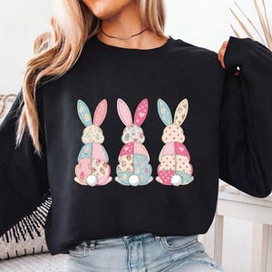 Peut inclure: Sweat-shirt noir à manches longues avec trois motifs de lapins en patchwork colorés. Chaque lapin présente des motifs floraux et géométriques roses, bleus et jaunes. Les lapins ont des queues touffues blanches et de longues oreilles.