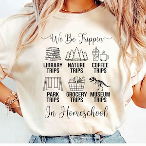 May include: A cream-coloured t-shirt featuring the phrase "We Be Trippin" and illustrations of books, trees, coffee, swings, a grocery basket, and a dinosaur. The text "In Homeschool" is also displayed.