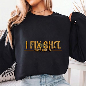 Op de afbeelding: Zwart sweatshirt met lange mouwen met de gele tekst "I FIX SHIT. THAT'S WHAT I DO". De tekst is gecentreerd en heeft een sleutel en een bladontwerp. De sweatshirt is gemaakt van zacht materiaal.