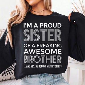May include: Black long-sleeve sweatshirt with the text "I'M A PROUD SISTER OF A FREAKING AWESOME BROTHER" in white. The text also includes "(...AND YES, HE BOUGHT ME THIS SHIRT)". The sweatshirt is worn by a person.