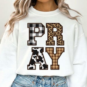 May include: White sweatshirt with the word "PRAY" in bold, decorative letters. The letter "P" is black and white checkered, "R" is leopard print, "A" is cow print, and "Y" is brown plaid. The sweatshirt is a casual style.