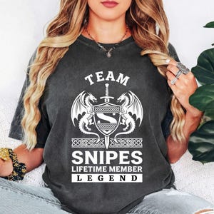 Peut inclure: T-shirt gris foncé avec un motif blanc. Le motif comprend un bouclier, des dragons et le texte "TEAM SNIPES LIFETIME MEMBER LEGEND". Le graphisme présente une épée et des éléments décoratifs, créant un thème médiéval ou fantastique.