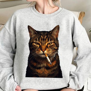 Pode incluir: Um moletom cinza claro com uma ilustração detalhada de um gato malhado marrom com um cigarro na boca. O gato tem uma expressão séria e a imagem é centralizada na frente do moletom.
