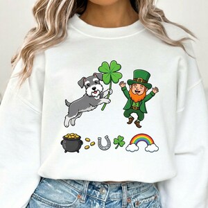 May include: White sweatshirt featuring a cartoon design with a gray dog holding a four-leaf clover, a leprechaun, a pot of gold, a horseshoe, a four-leaf clover, and a rainbow. The design is in a cartoon style.