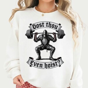 May include: White sweatshirt featuring a graphic of a knight lifting a barbell. The knight is clad in silver armor and is in a squat position. The text "Dost thou" is above the knight, and "Even hoist?" is below.