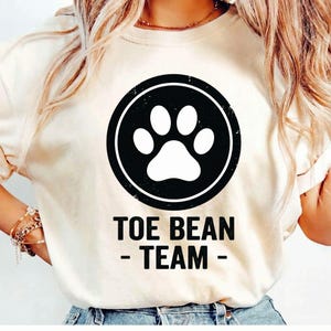 May include: Cream-colored t-shirt featuring a black circle with a white paw print in the center. The words "TOE BEAN - TEAM -" are printed in black below the circle. The shirt is made of a soft material.