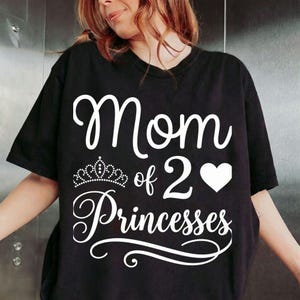 Peut inclure: T-shirt noir avec l'inscription blanche "Mom of 2 Princesses". Une couronne est au-dessus du mot "Princesses", et un cœur blanc est à côté du chiffre "2". Le t-shirt est de style décontracté.