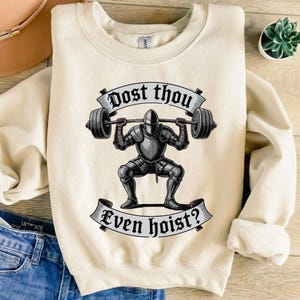 May include: Cream-colored sweatshirt with a graphic of a knight lifting weights. The knight is in full armor, and the text "Dost thou Even hoist?" is displayed above and below the knight in a banner.
